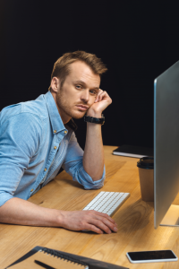Un homme, l'air las et pensif, assis devant un écran d'ordinateur sur un bureau en bois, la main sous le menton, symbolisant l'incertitude ou la lassitude professionnelle dans le secteur IT