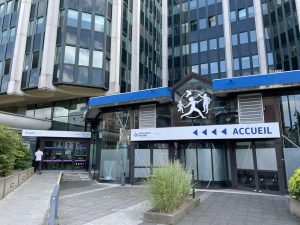 Façade du bâtiment d'accueil de la CPAM (Caisse Primaire d'Assurance Maladie) à Lille, avec enseigne "L'Assurance Maladie", "ACCUEIL", et logo d'une famille stylisée au-dessus de l'entrée. Un grand immeuble de bureaux vitré se dresse derrière.